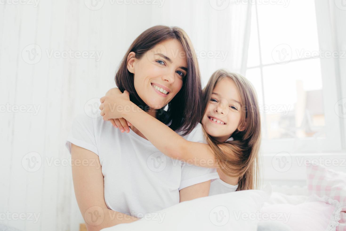 toma interior de una guapa madre morena con una sonrisa amable y su pequeña hija le da un abrazo, disfruta del ambiente doméstico, posa contra el espacioso interior del dormitorio blanco. concepto de maternidad y familia foto