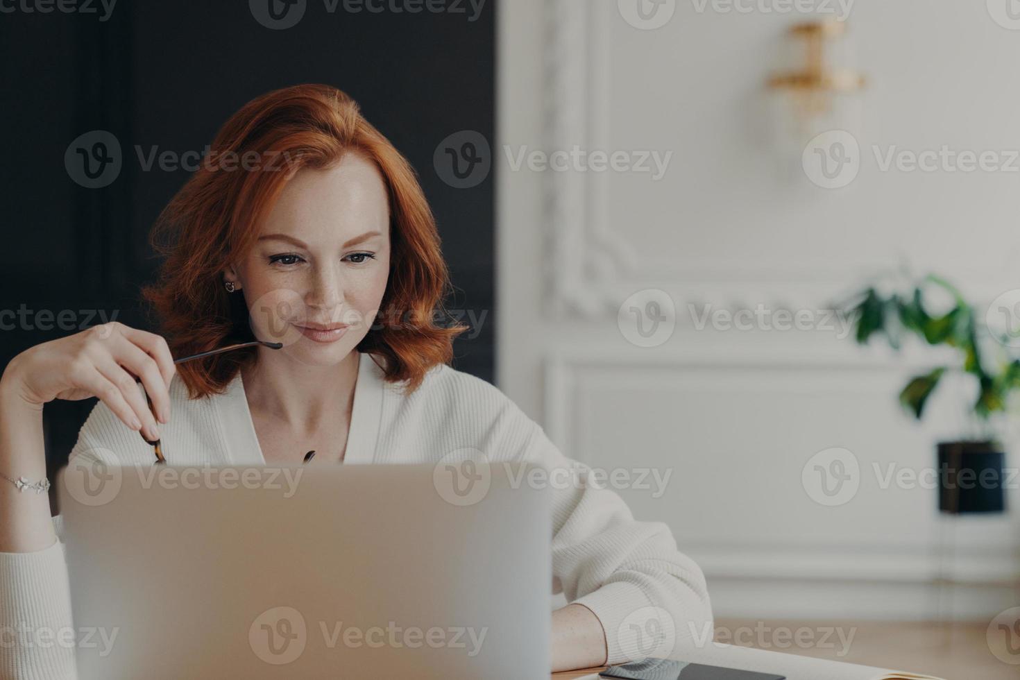 Photo of serious redhead woman journalist concentrated at laptop computer, searches information for article on internet website, holds spectacles, wears white jumper, checks email, works remotely