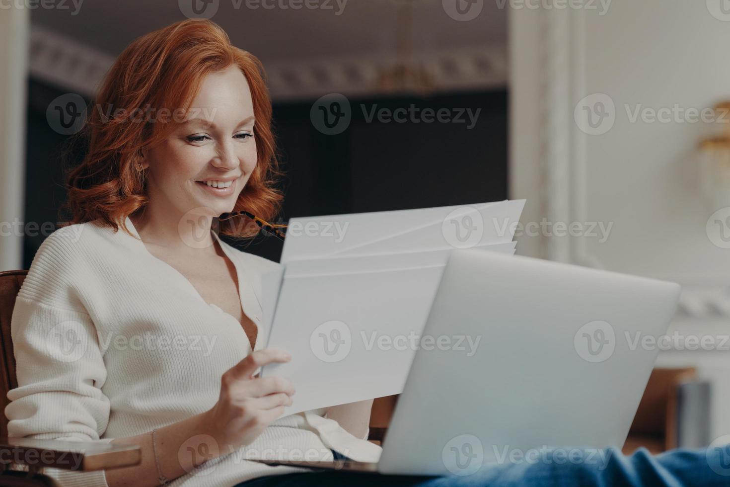 Horizontal view of pleased adult woman entrepreneur works from home, works remotely from home, looks through paper documents, uses laptop computer, wears casual jumper, poses in own cabinet. photo