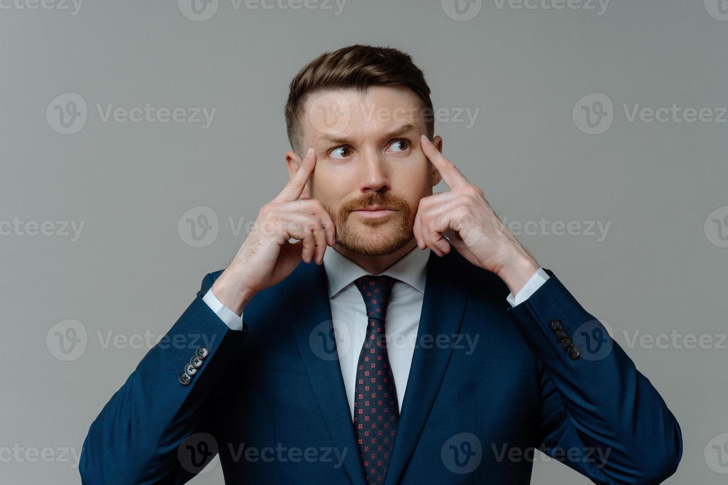 Indoor shot of motivated thoughtful bearded businessman keeps fingers on temples looks away gets ready for conference dressed in formal clothes isolated over grey background has hard working day photo