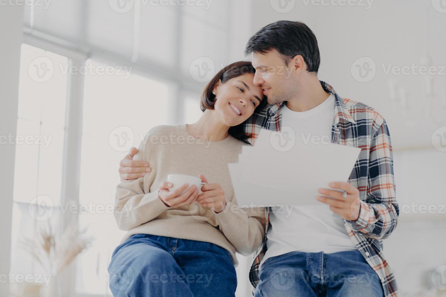 Indoor shot of relaxed beautiful woman with coffee cup, receives hug from husband who holds papers, make financial report together, own business company. Family, finances and budget concept. photo