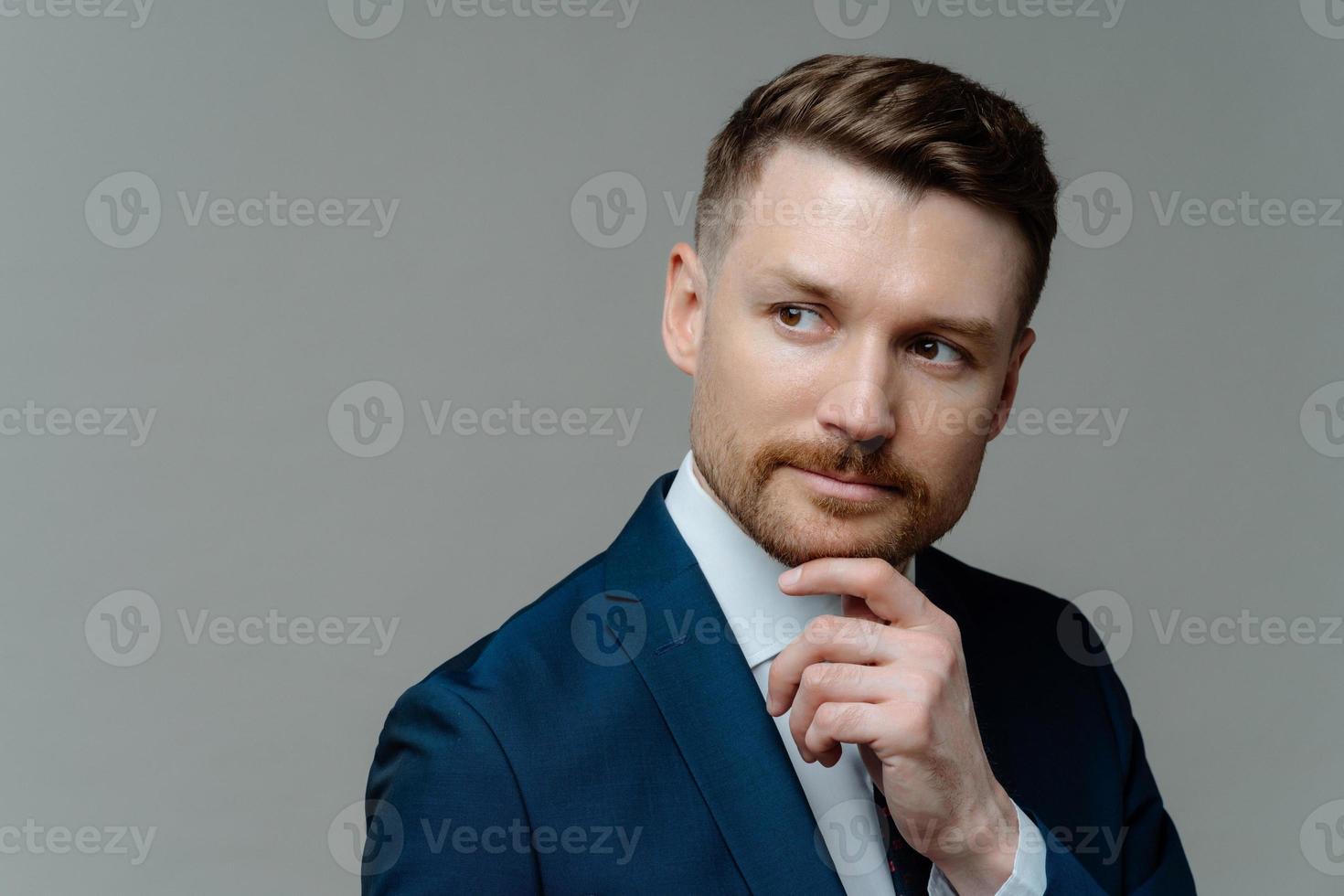 Thoughtful bearded businessman holds chin considers something looks away makes decision dressed in formal clothes looks forward future opportunity isolated over grey background blank space for text photo