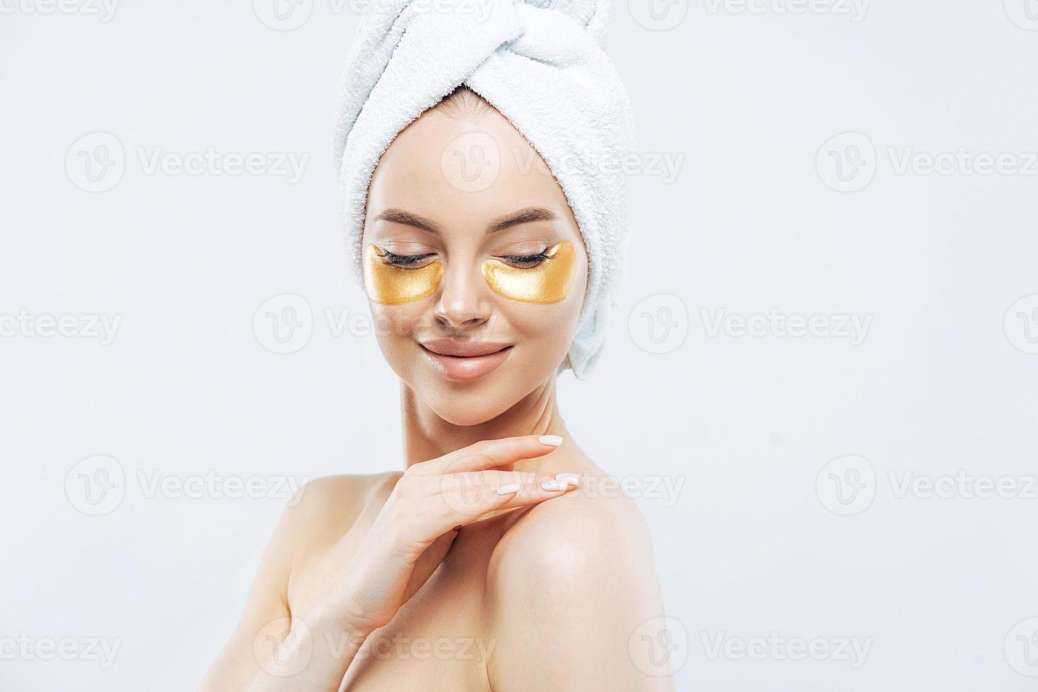 Sideways shot of tender young woman with eye collagen gold pads, healthy fresh skin, anti aging moisturising mask, touches shoulder gently, wears bath towel on head, isolated over white studio wall photo