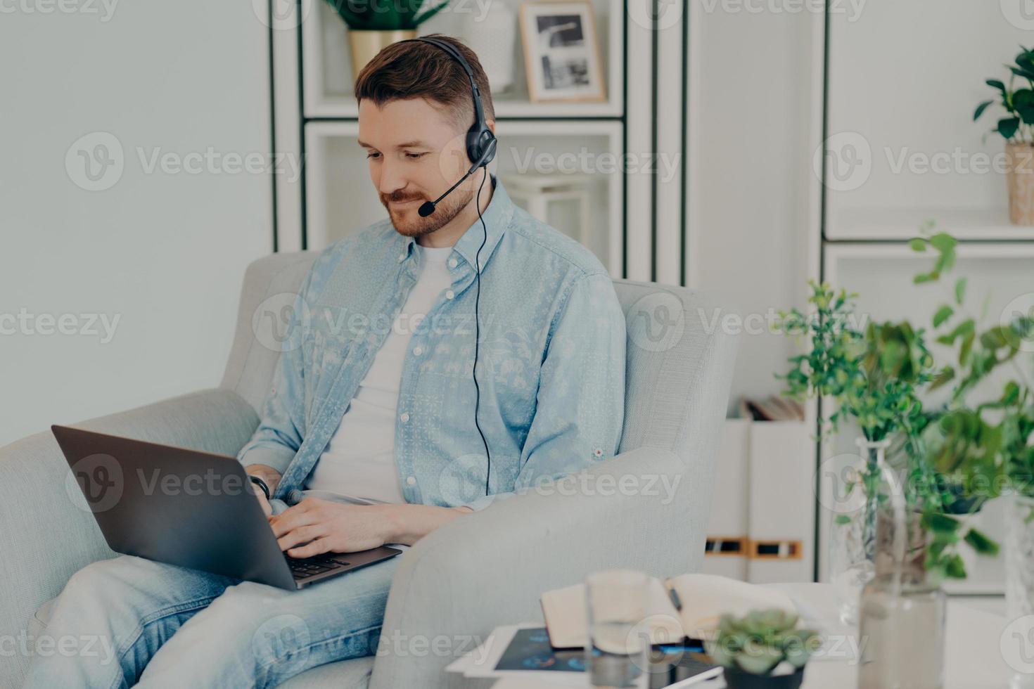 Busy man dressed in denim clothes supplies consultant service types on laptop computer wears headphones with microphone works distantly sits in comfortable armchair participates in online negotiations photo