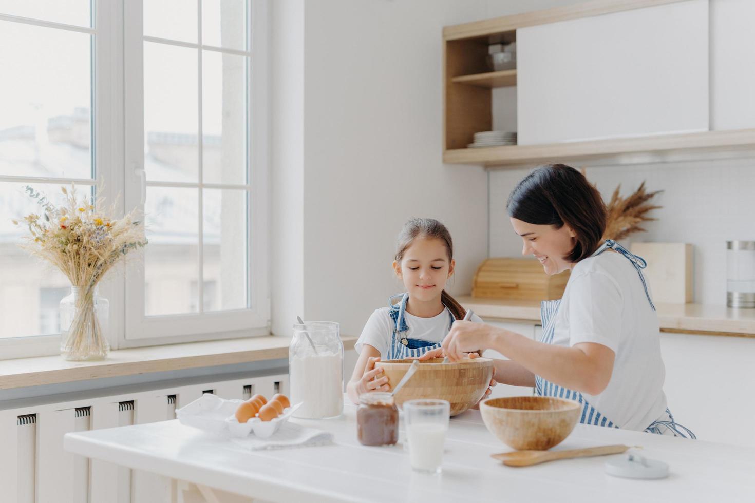 Brunette woman with smile shows little daughter how to cook, gives culinary lesson, bought different products in shop for cooking, wear aprons, smile pleasantly, window with vase, kitchen interior photo