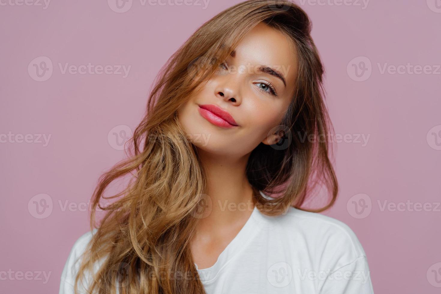 Portrait of satisfied relaxed young female model tilts head, has makeup, fair hair, dressed in white clothes, poses against purple background, has well cared complexion. People, beauty, face care photo