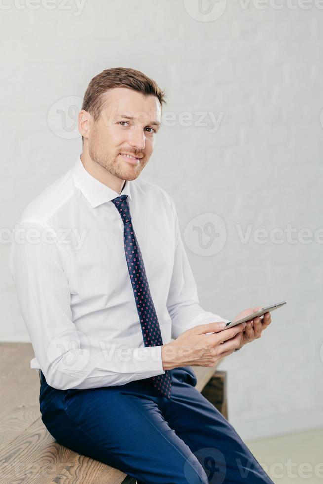 Confident pleased young male entrepreneur in luxury formal clothes, reads recieved text message from colleague on modern tablet computer, looks joyfully at camera, poses in office. Vertical shot photo