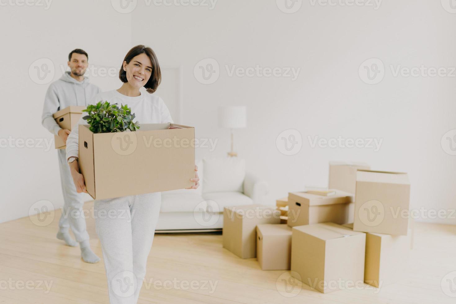 Happy woman and her husband carry boxes with personal belongings, being busy during relocation in other place for living, enter new home, move together in big house, unpack household things. photo