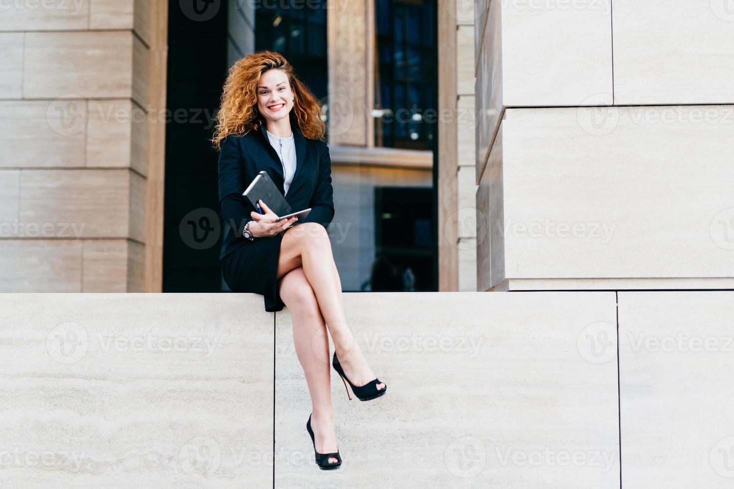 joven atractiva con el pelo rizado, vestida con traje formal negro y zapatos con tacones altos, sosteniendo su cuaderno y tableta, haciendo arreglos con sus socios comerciales, con buen humor foto