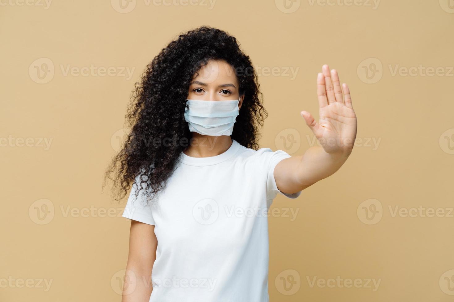 Serious dark skinned woman makes stop gesture, pulls palm towards camera, wears medical flu mask, asks stay at home not to spread coronavirus disease, prevents virus, dressed in white t shirt. photo