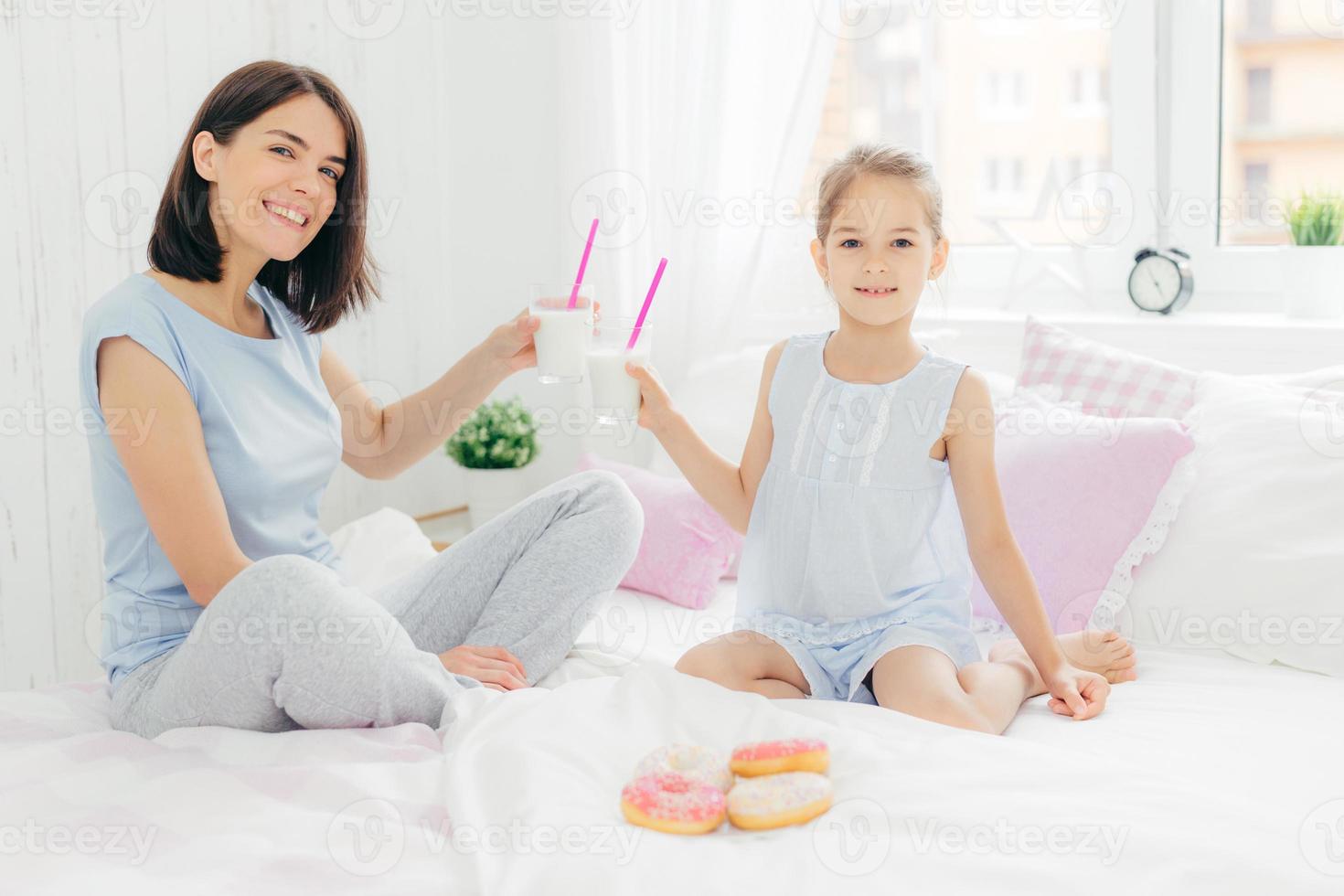 retrato de una joven madre feliz y su pequeña hija sostienen vasos de batido de leche, desayunan en la cama, miran positivamente a la cámara, disfrutan el fin de semana en casa y juntos, vestidos con ropa de dormir foto