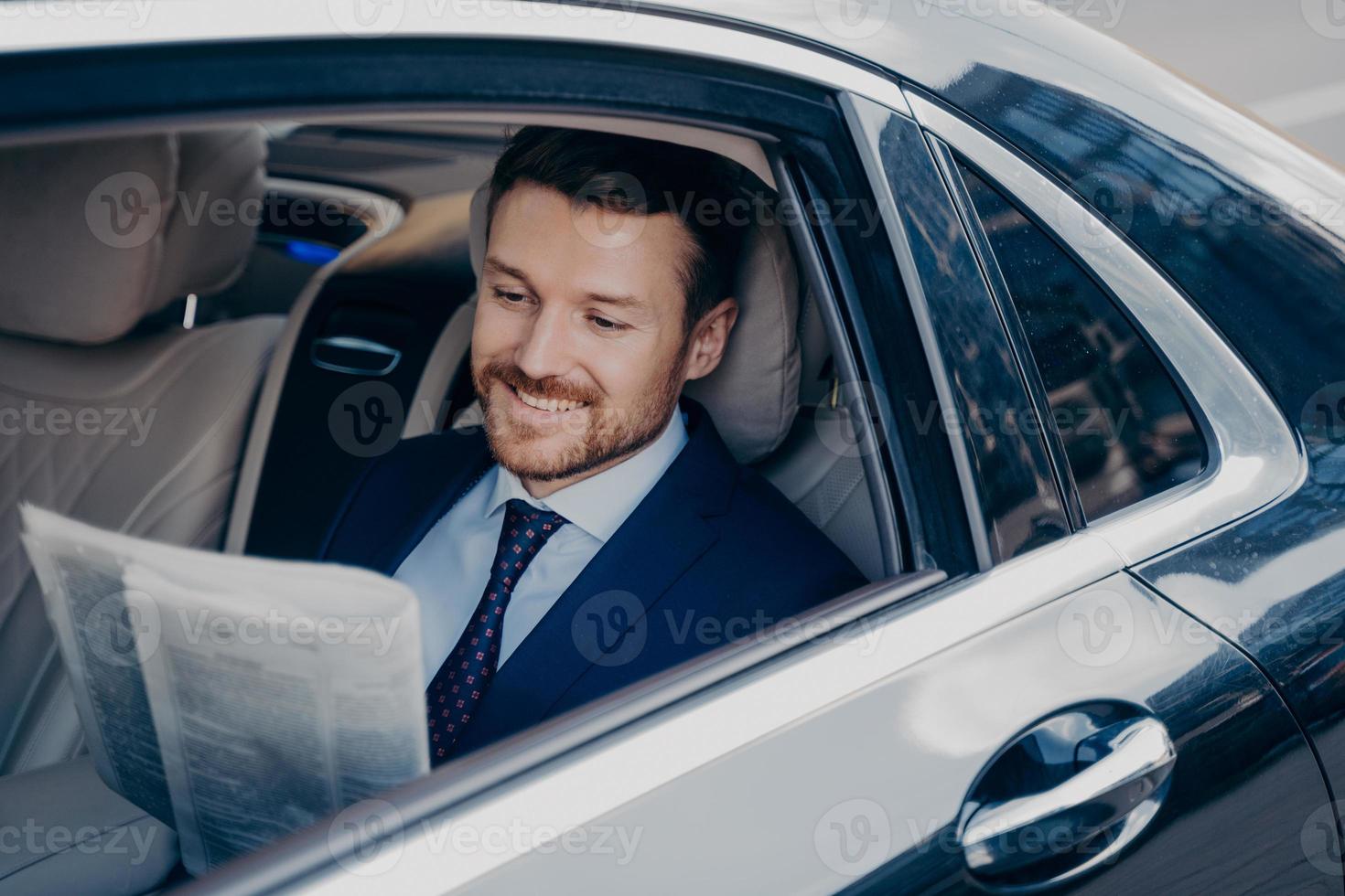 apuesto joven banquero con elegante traje formal de esmoquin lee el periódico en un auto de lujo foto