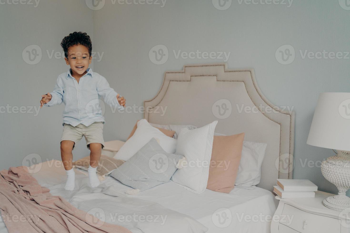 Happy carefree mixed race ethnicity kid boy in casual clothes having fun and jumping on bed photo