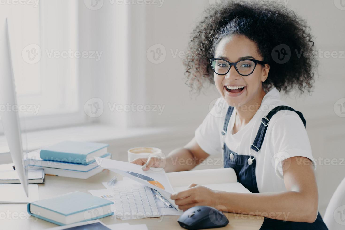 una foto lateral de una feliz y próspera empresaria sostiene papel, estudia un gráfico, hace planes, está ocupada con el papeleo, se sienta en un escritorio de madera con una computadora, usa anteojos, camiseta y overoles