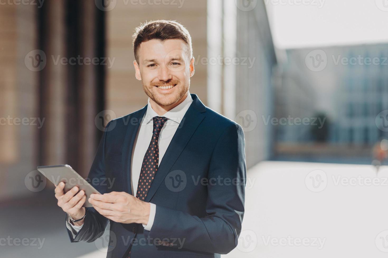 Portrait of handsome male with positive expression, updates software on tablet computer, connected to wireless internet, shares files during remote job, stands outdoor, reads companys news. photo