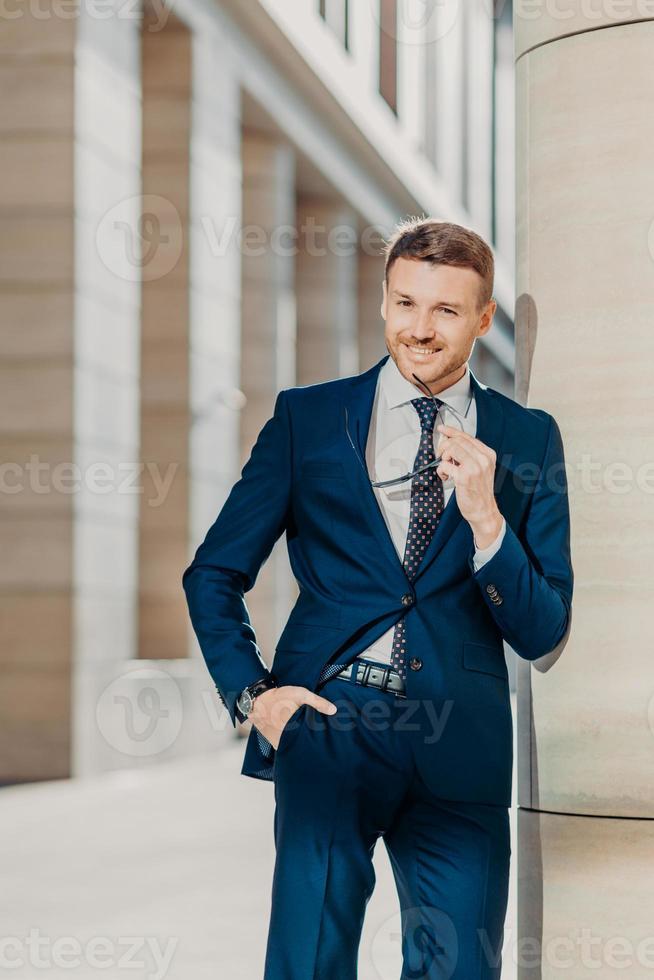 Thoughtful cheerful bearded male entrepreneur has pleasant smile, wears black suit, holds sunglasses, poses indoor, being delighted with successful business meeting and signing contract with partner photo