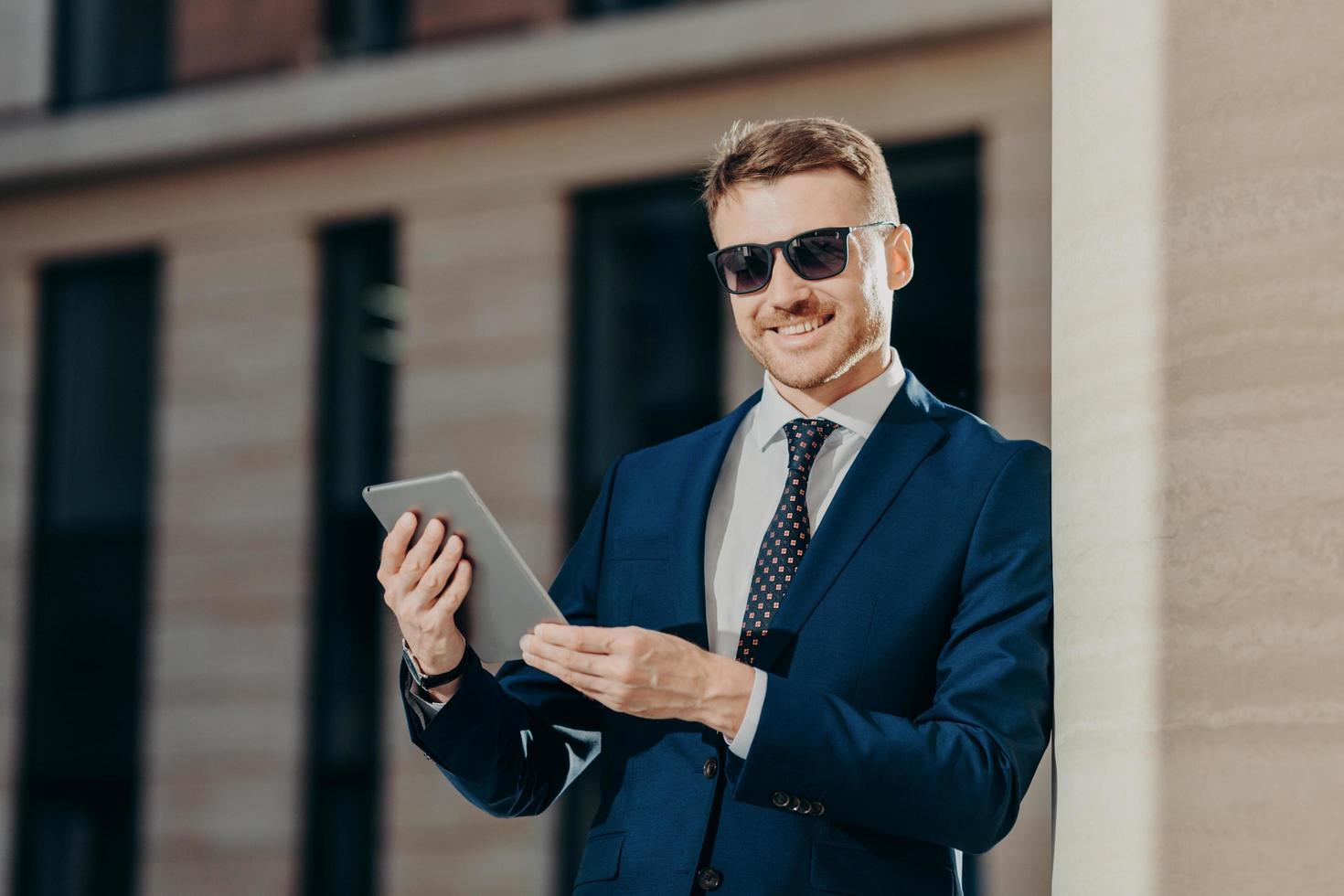 Portrait of happy male entrepreneur sends text message to colleague via touch pad, connected to wireless internet, updates software, wears trendy sunglasses and black suit. People and technology photo