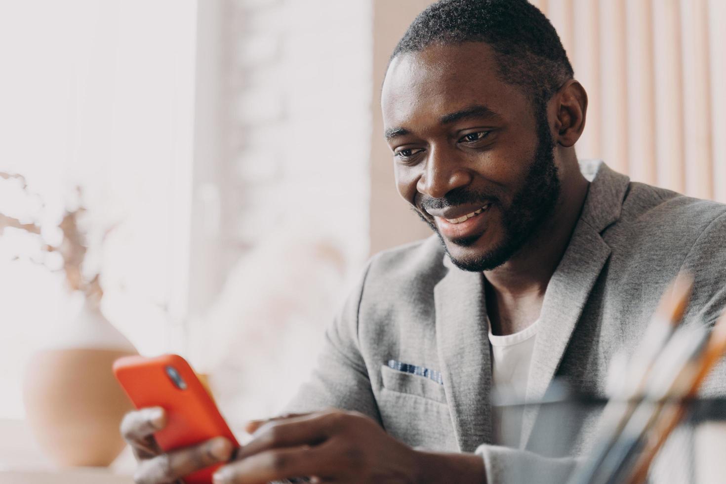 Smiling african american businessman using mobile, chatting online while sitting at office desk photo