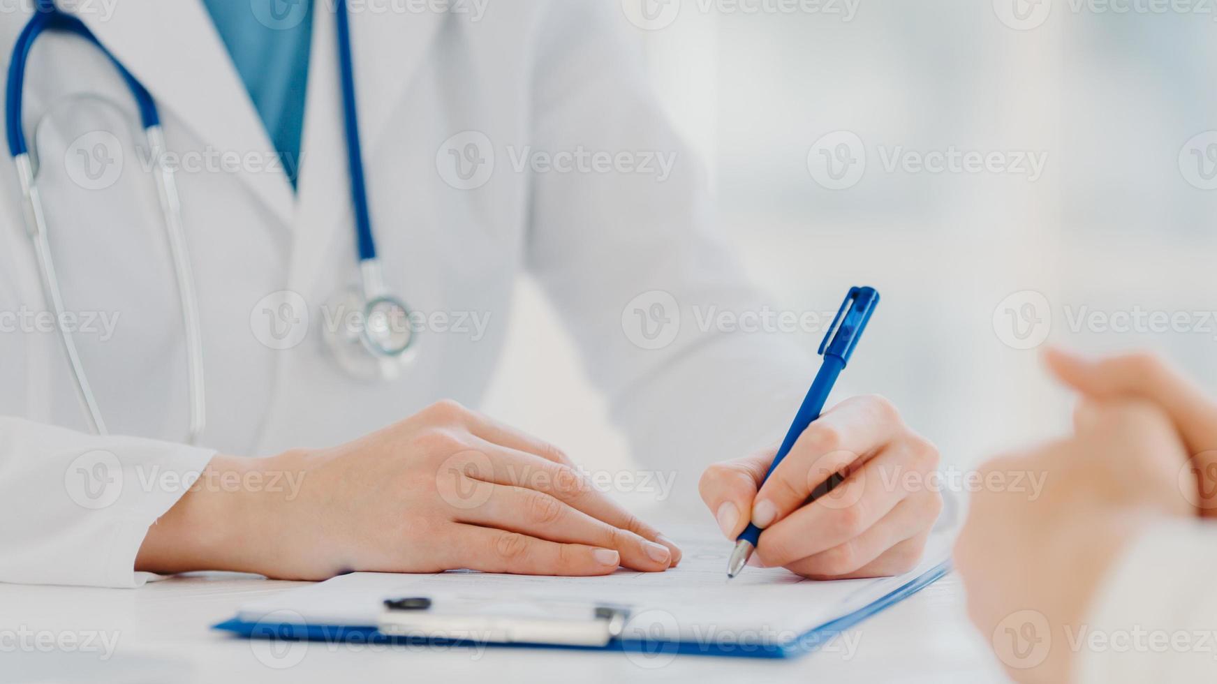 Unrecognizable woman doctor writes down in clipboard, makes prescription for patient, records data for analysis, wears white gown. Close up, focus on hands. Medicine, insurance, health care concept photo