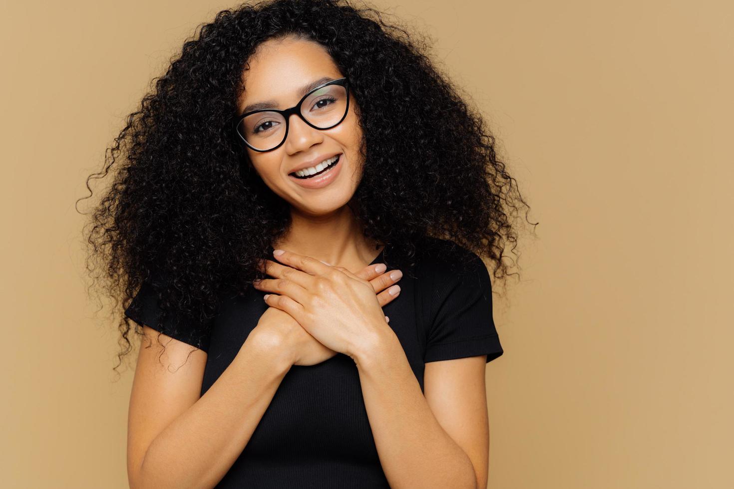 Touched lovely woman with curly hairstyle, keeps palms on chest, tilts head, has charming smile on face, wears optical glasses, black casual t shirt, models against brown background. Gratitude photo