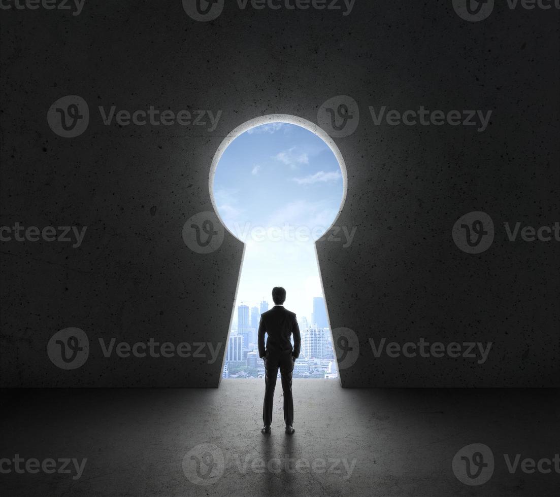 Businessman standing in front of bright big keyhole door, in a concrete wall with a view to the city photo