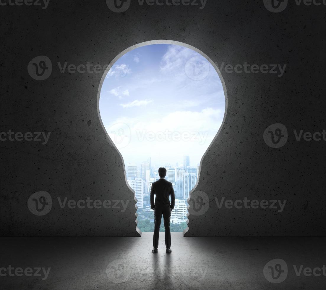 Businessman standing in front of bright big light bulb shining door in a concrete wall with a view to the city photo