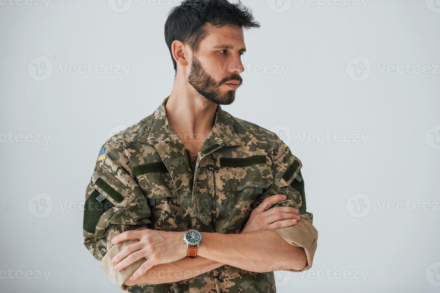 Arms crossed. Soldier in uniform is standing indoors against white wall photo