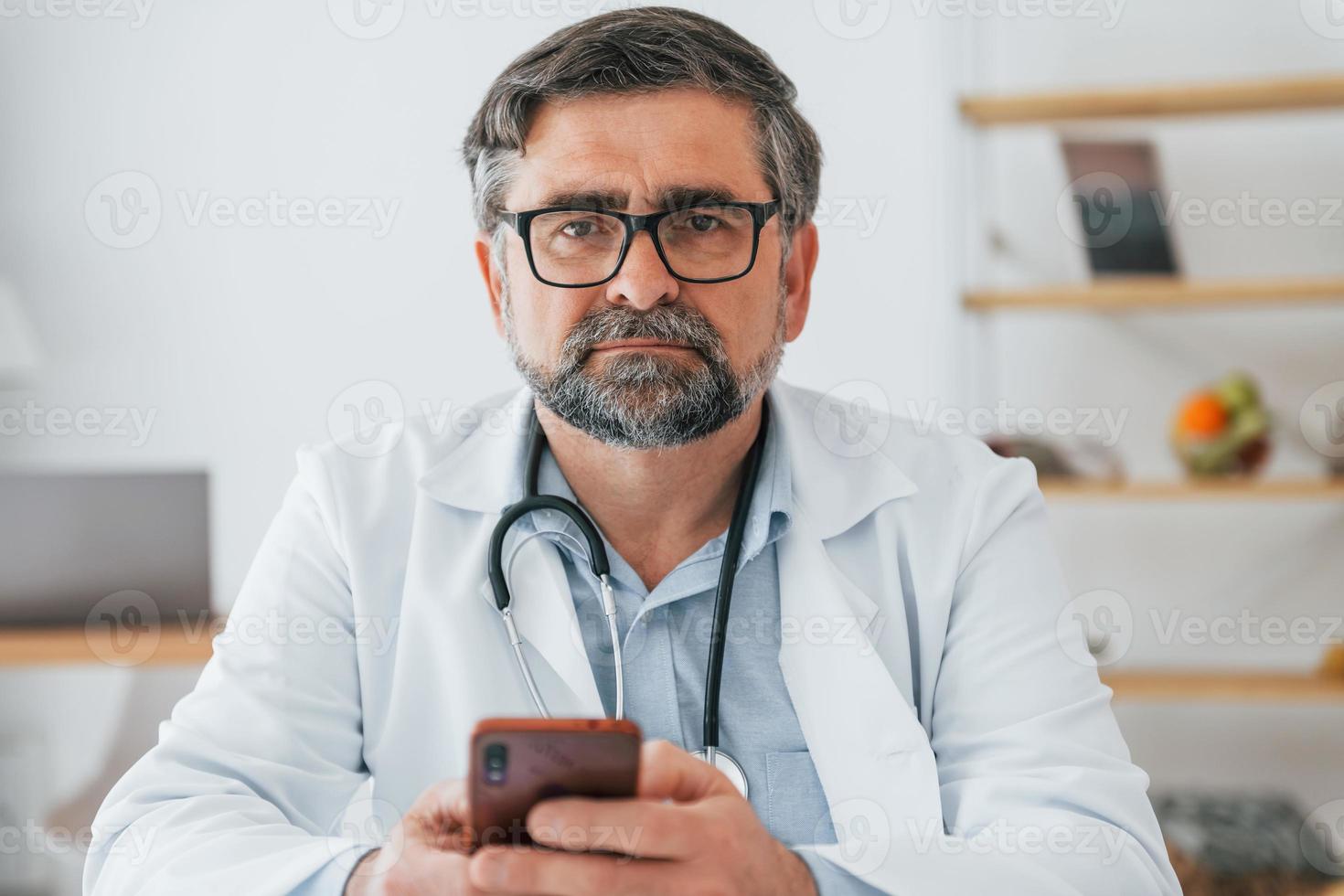 Male doctor is sitting with phone in hands. Professional medical worker in white coat is in the office photo
