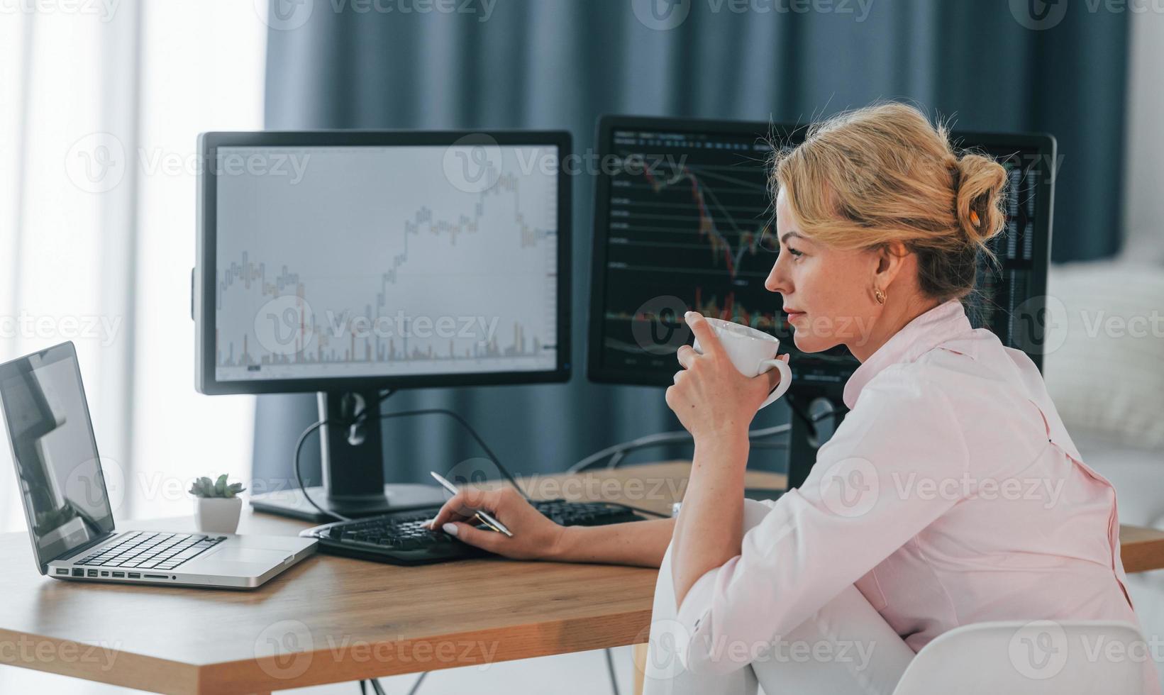 Sitting with cup of drink in hands and monitoring market. Female stock broker in formal wear is working in the office photo