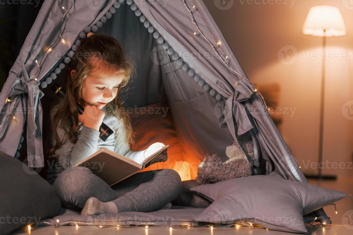 Reading book at evening. Cute little girl playing in the tent that is in the domestic room photo