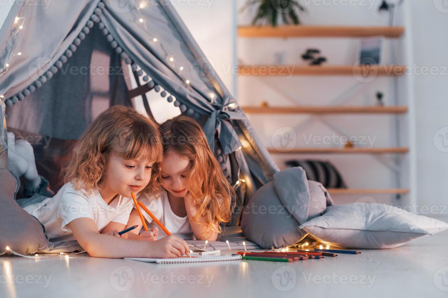 Learning to write and draw. With paper and pencils. Two little girls is in the tent in domestic room together photo