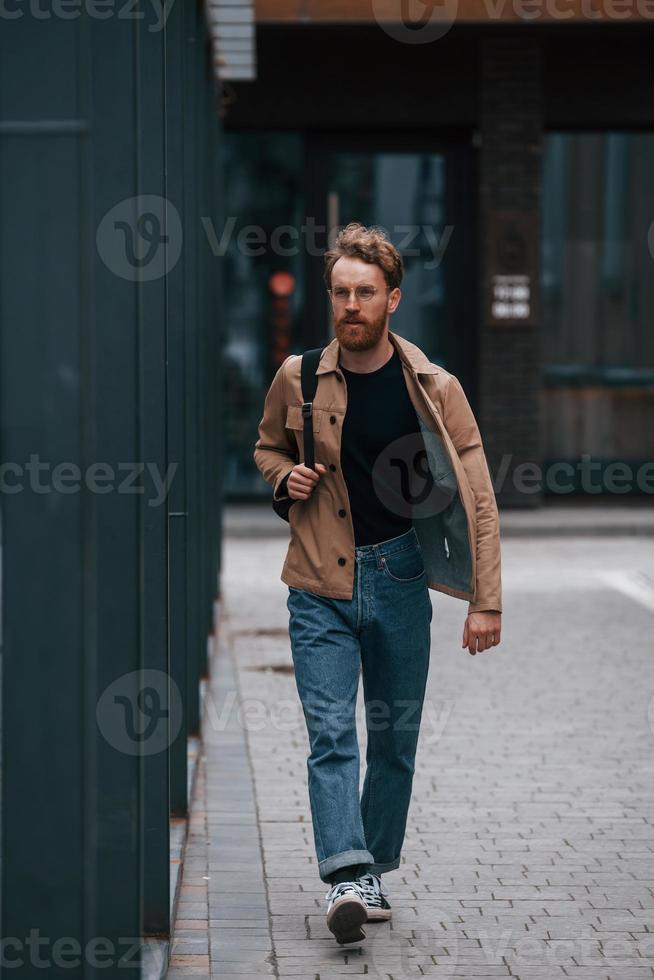 Front view. Stylish man with beard in khaki colored jacket and in jeans is outdoors near building photo