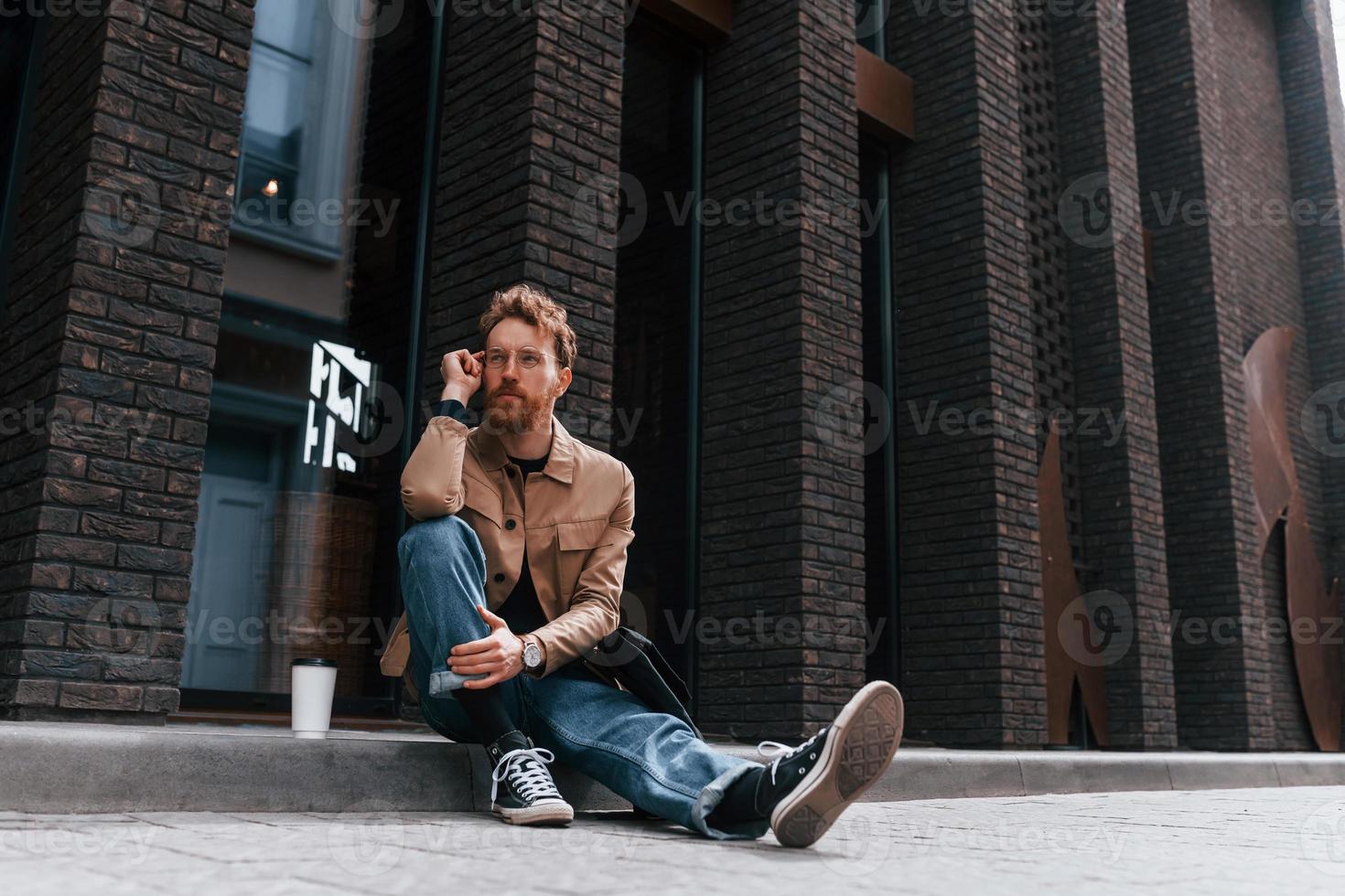 Coffee cup. Stylish man with beard in khaki colored jacket and in jeans is outdoors near building photo