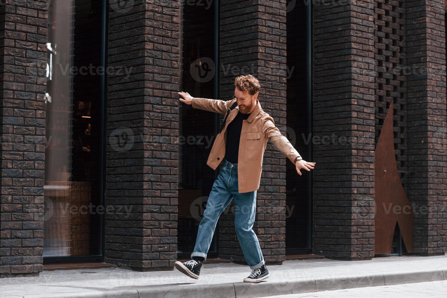 Having fun by walking the sidewalk. Stylish man with beard in khaki colored jacket and in jeans is outdoors near building photo