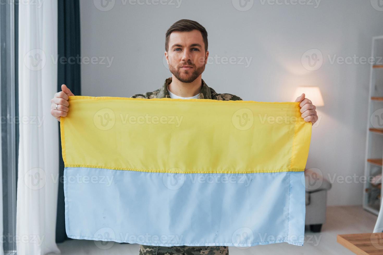 Soldier in uniform holding Ukrainian flag in hands at home photo
