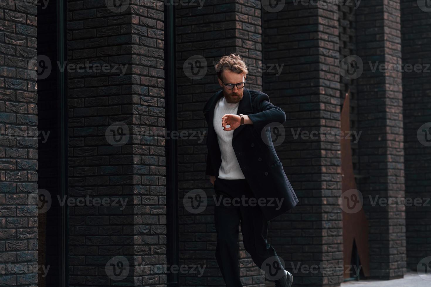 Running out of time. Stylish man with beard and in glasses is outdoors near building photo