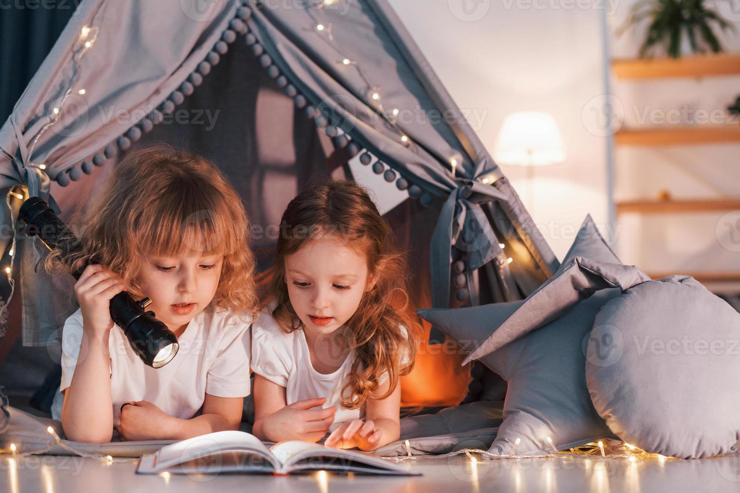 Reading book and using flashlight. Two little girls is in the tent in domestic room together photo