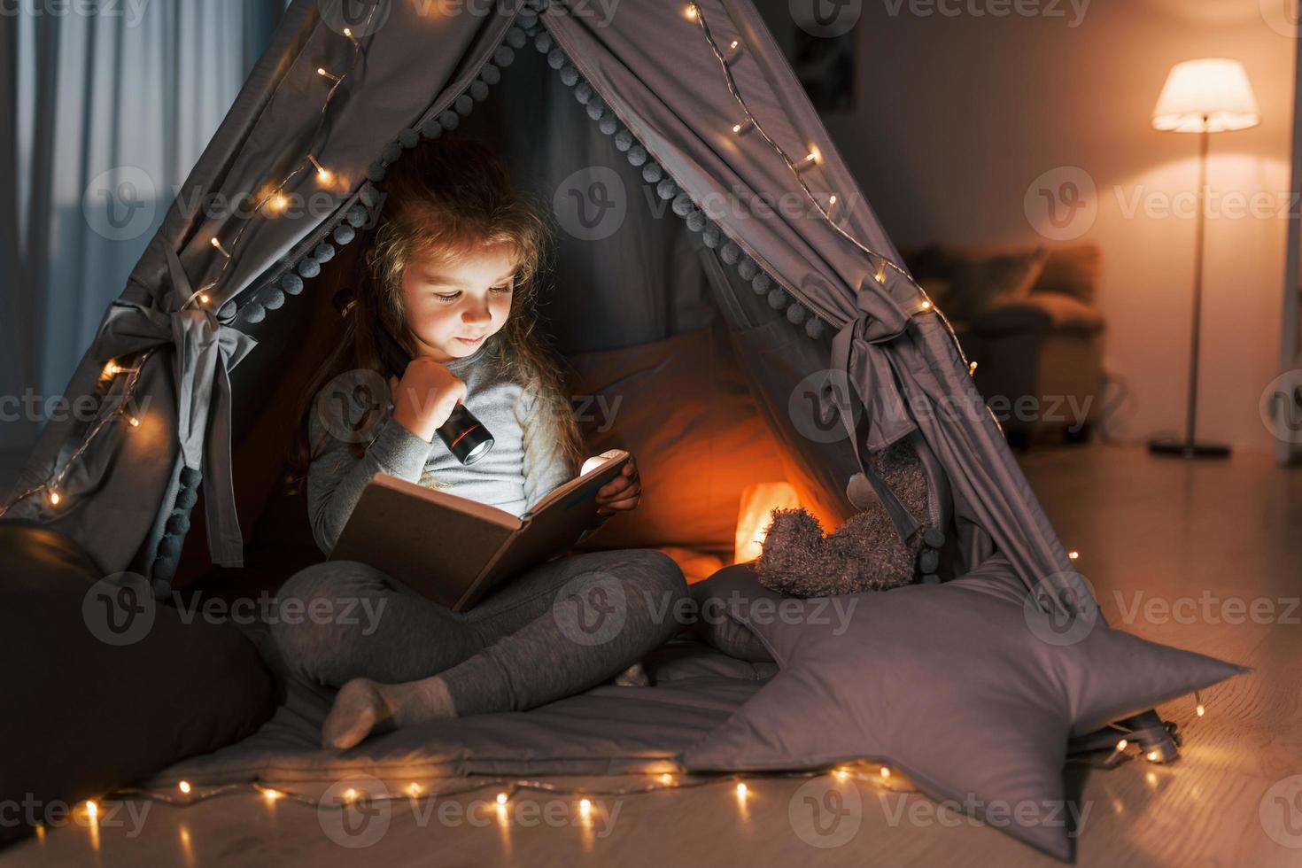 Reading book at evening. Cute little girl playing in the tent that is in the domestic room photo