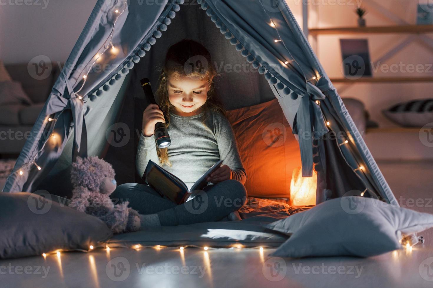 Using flashlight to read the book. Cute little girl playing in the tent that is in the domestic room photo