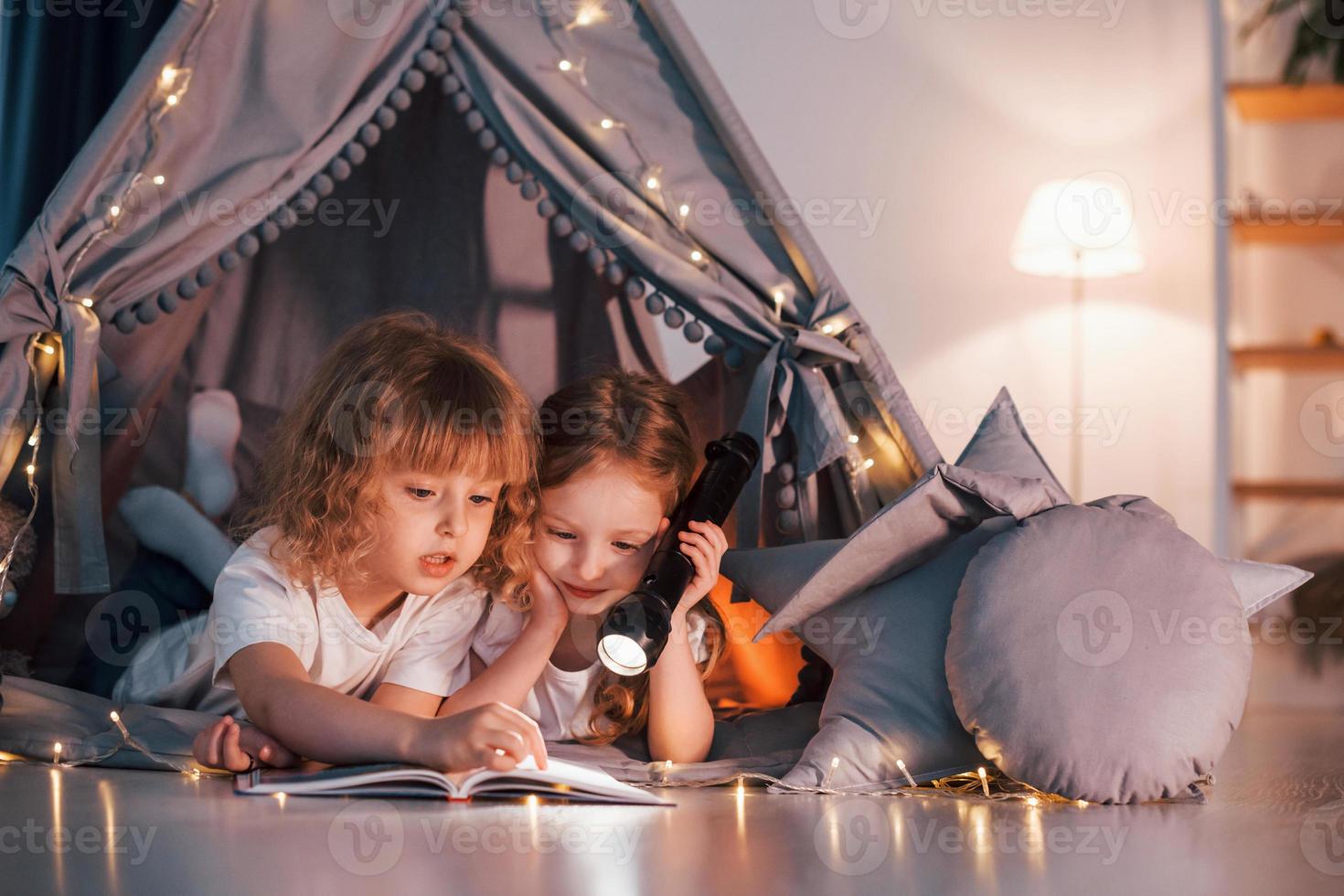 Laying down and using flashlight to read the book. Two little girls is in the tent in domestic room together photo