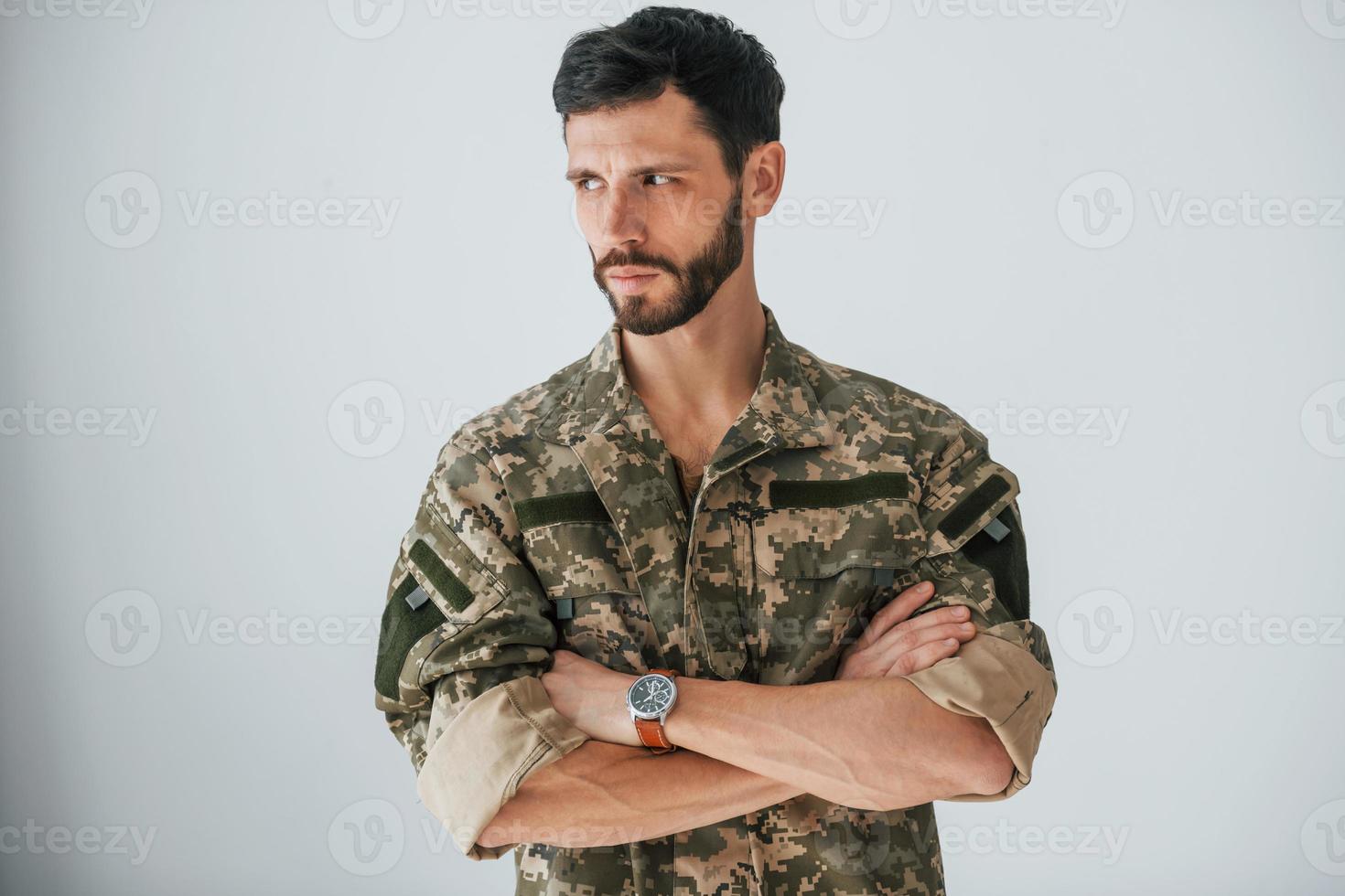 Arms crossed. Soldier in uniform is standing indoors against white wall photo