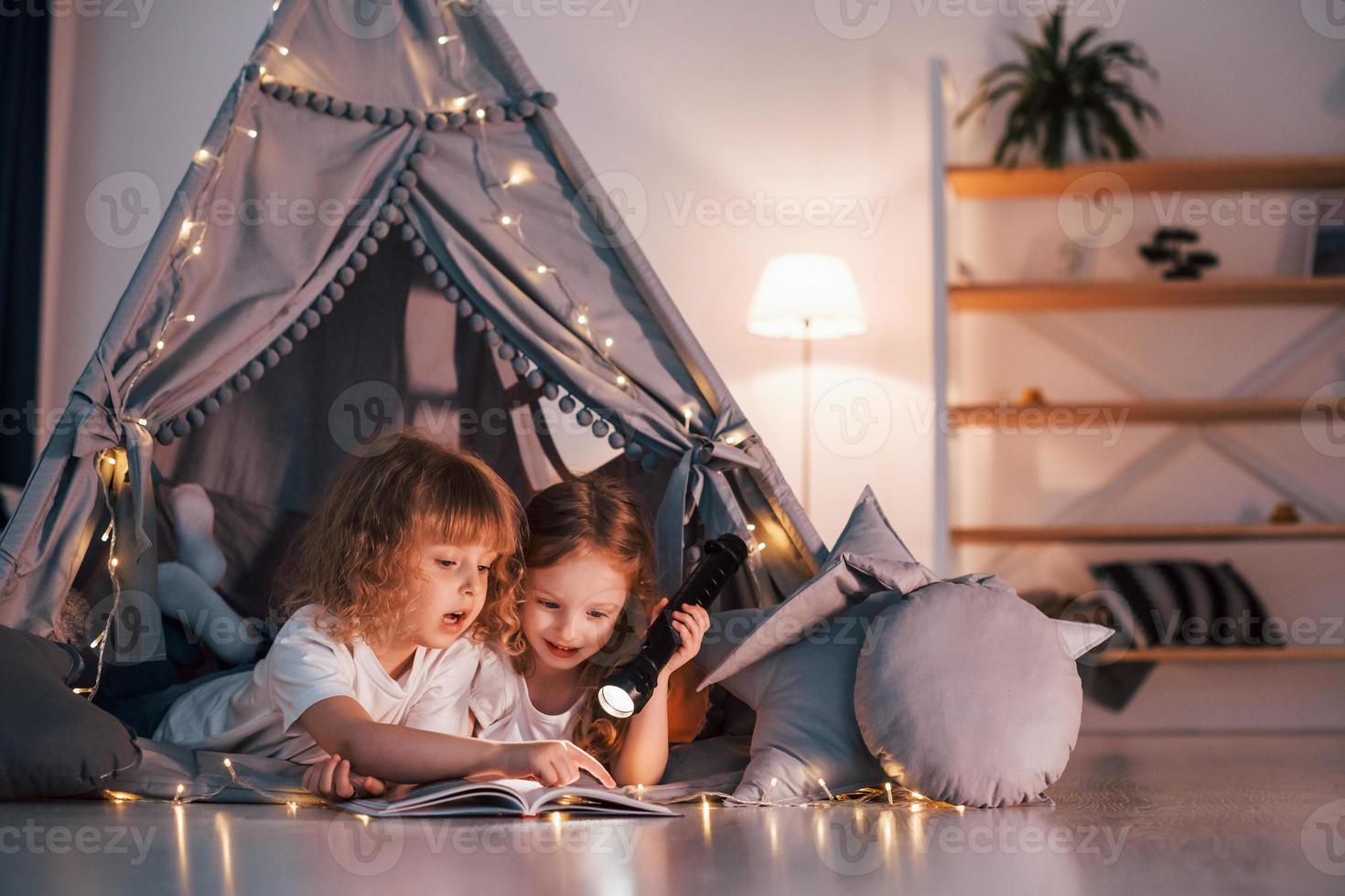Laying down and using flashlight to read the book. Two little girls is in the tent in domestic room together photo