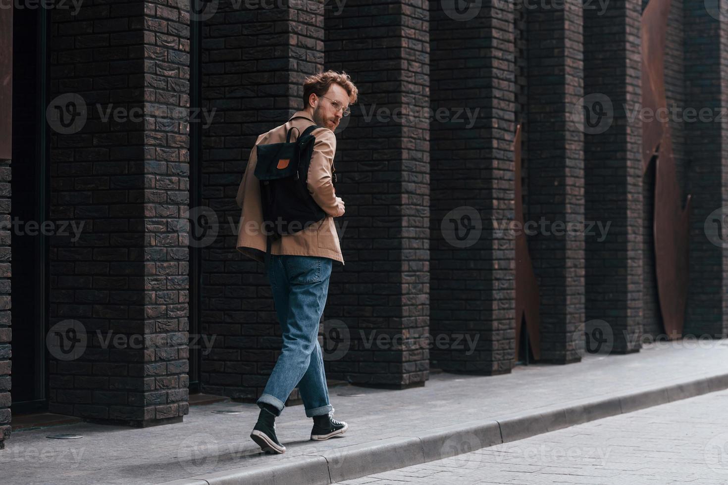Stylish man with beard in khaki colored jacket and in jeans is outdoors near building photo