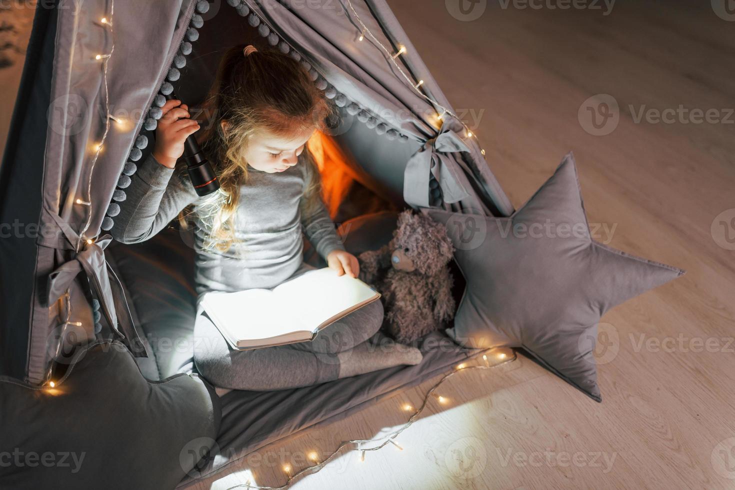 Reading book at evening. Cute little girl playing in the tent that is in the domestic room photo