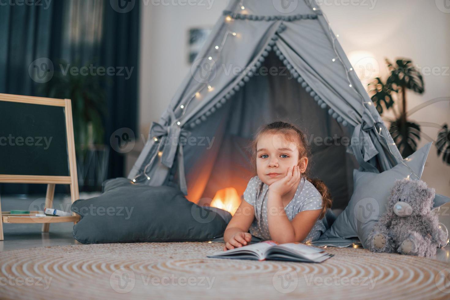 Reading the book. Cute little girl playing in the tent that is in the domestic room photo