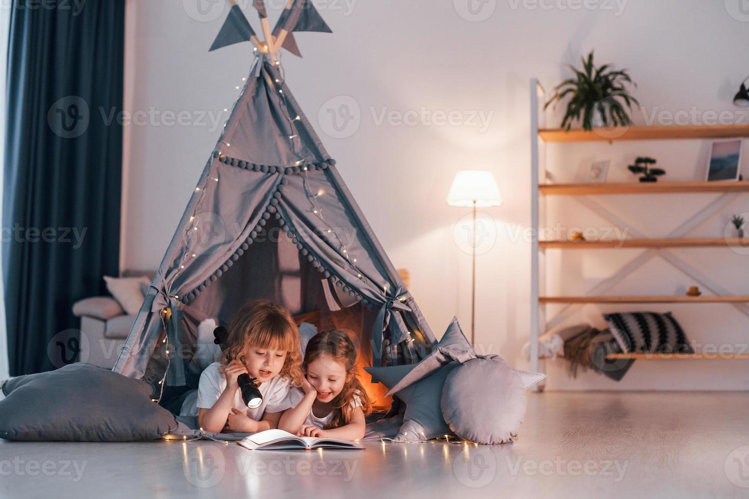 Laying down and using flashlight to read the book. Two little girls is in the tent in domestic room together photo