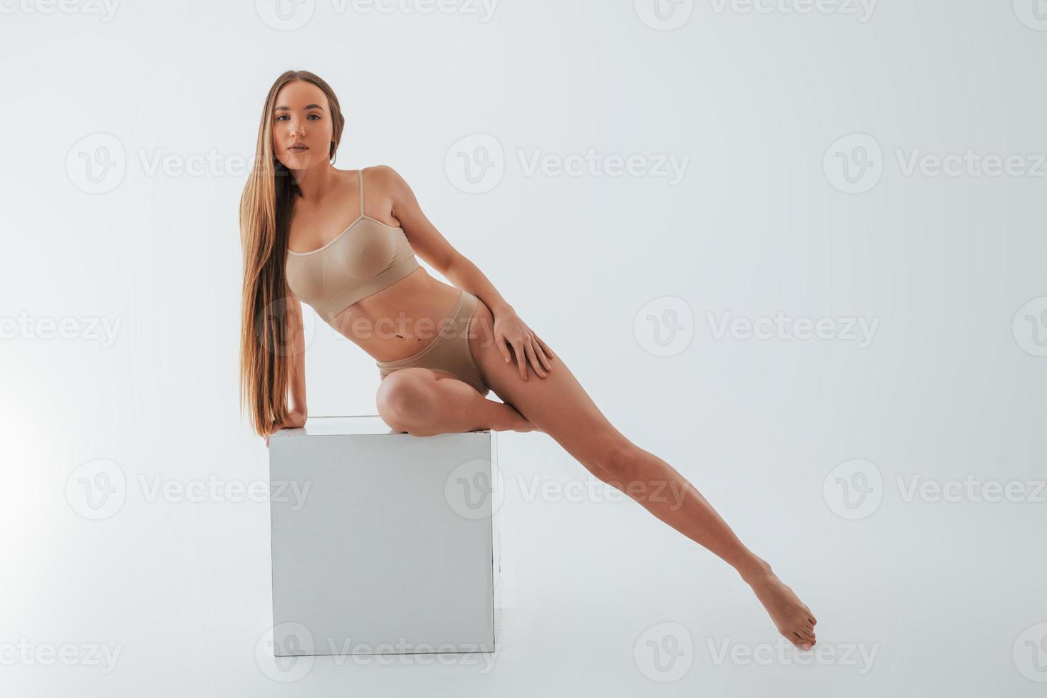 Sitting on the one leg on the white cube. Woman in underwear with slim body type is posing in the studio photo