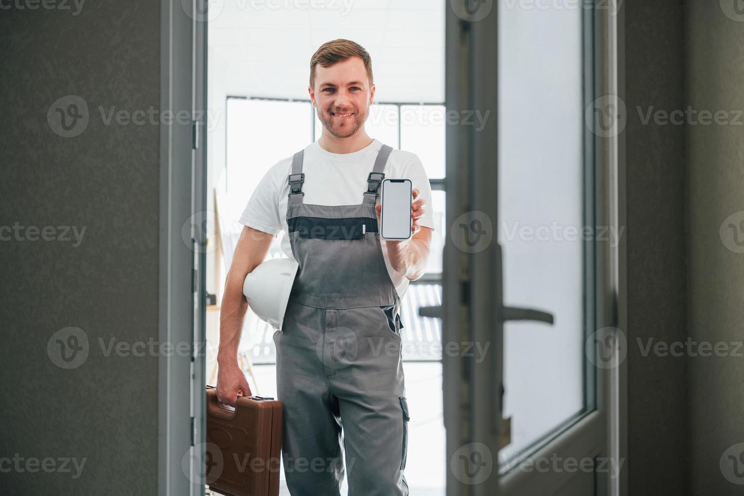 With smartphone in hand. Repairman is standing indoors in the modern room photo