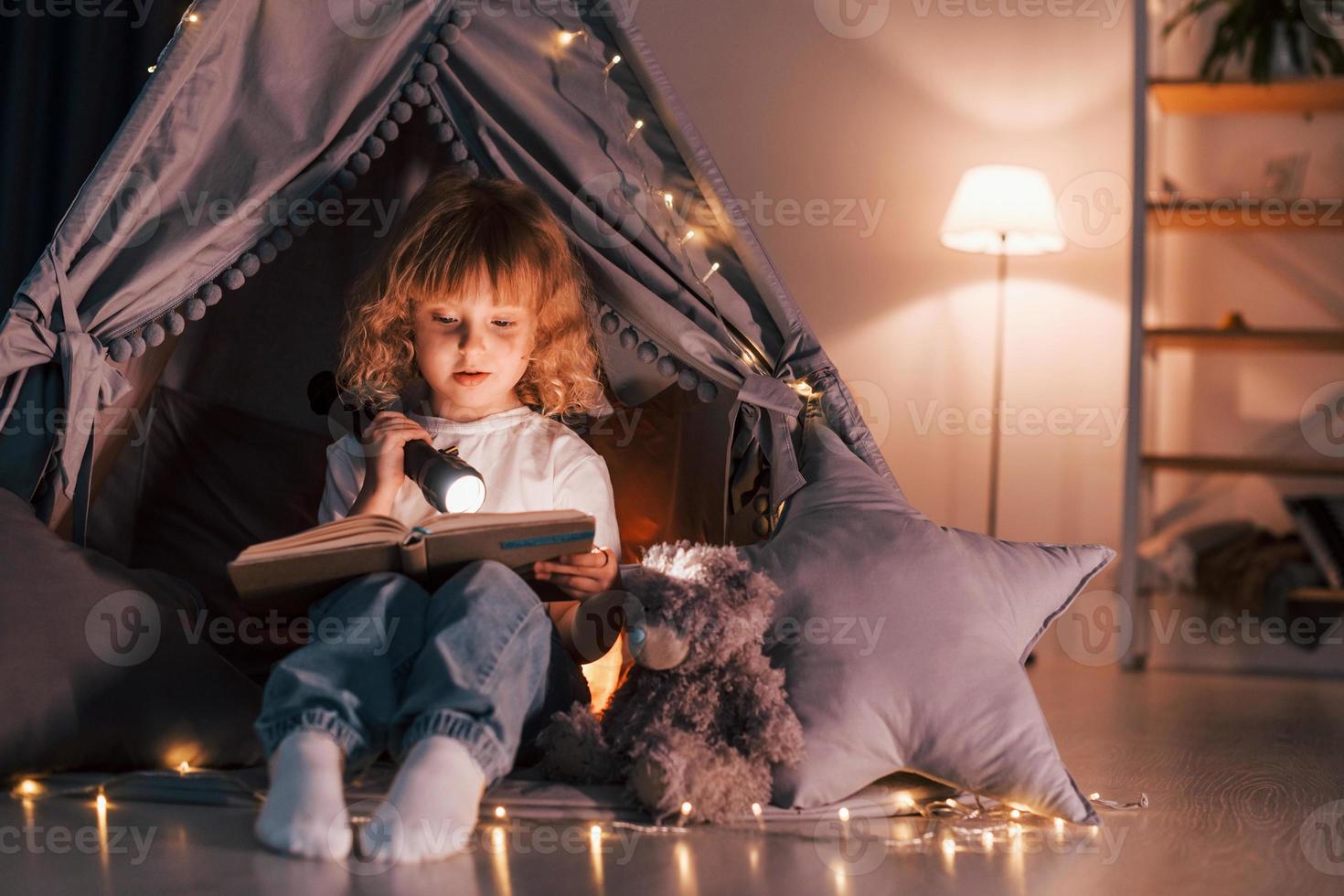 Using flashlight to read the book. Cute little girl is in the tent in domestic room photo