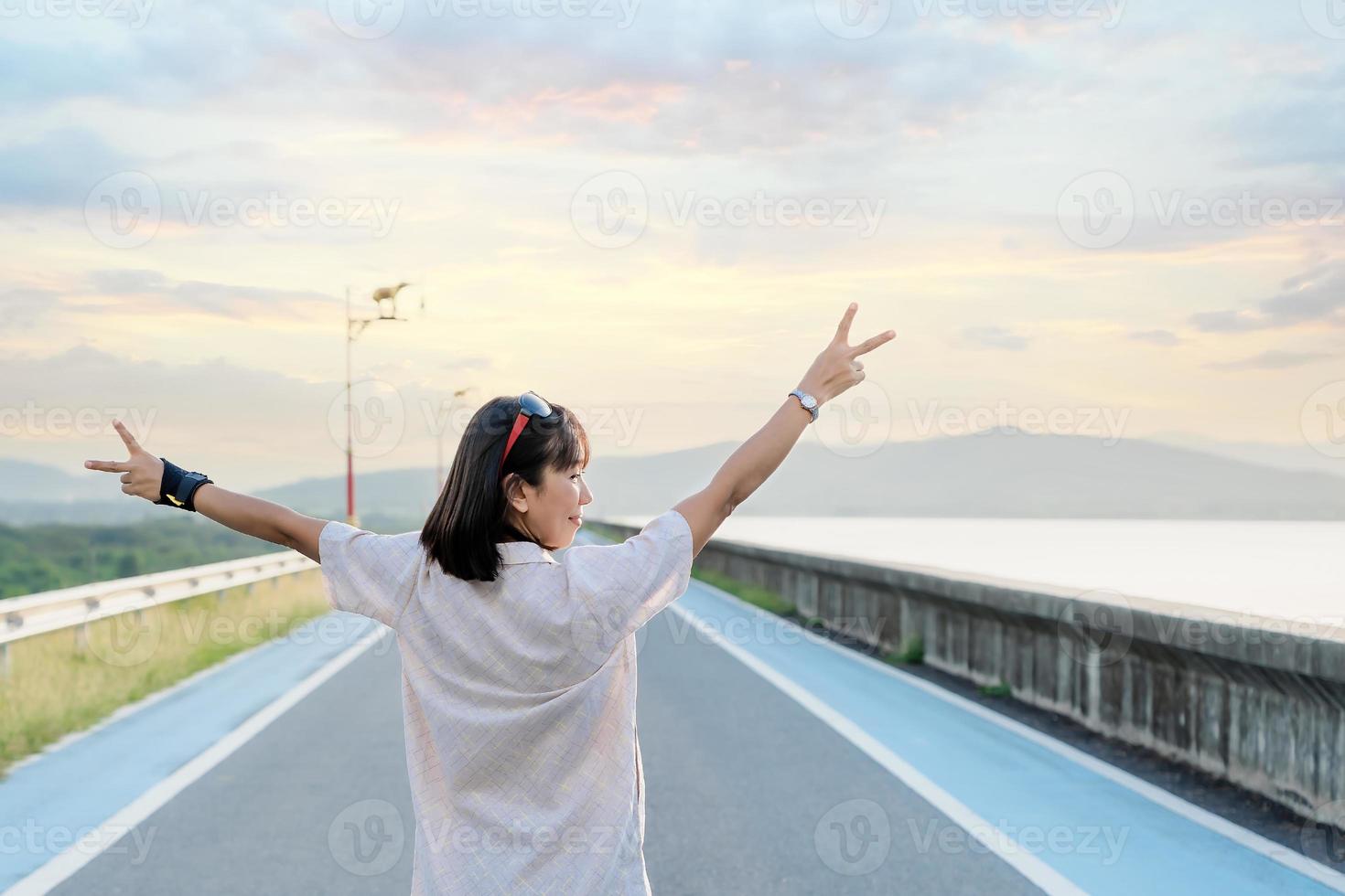 Back view of a female tourist rising hands up in the air. Woman traveler standing on street of dam with a sunset background on a vacation trip. Asian lady enjoying nature on holiday. Freedom concept photo